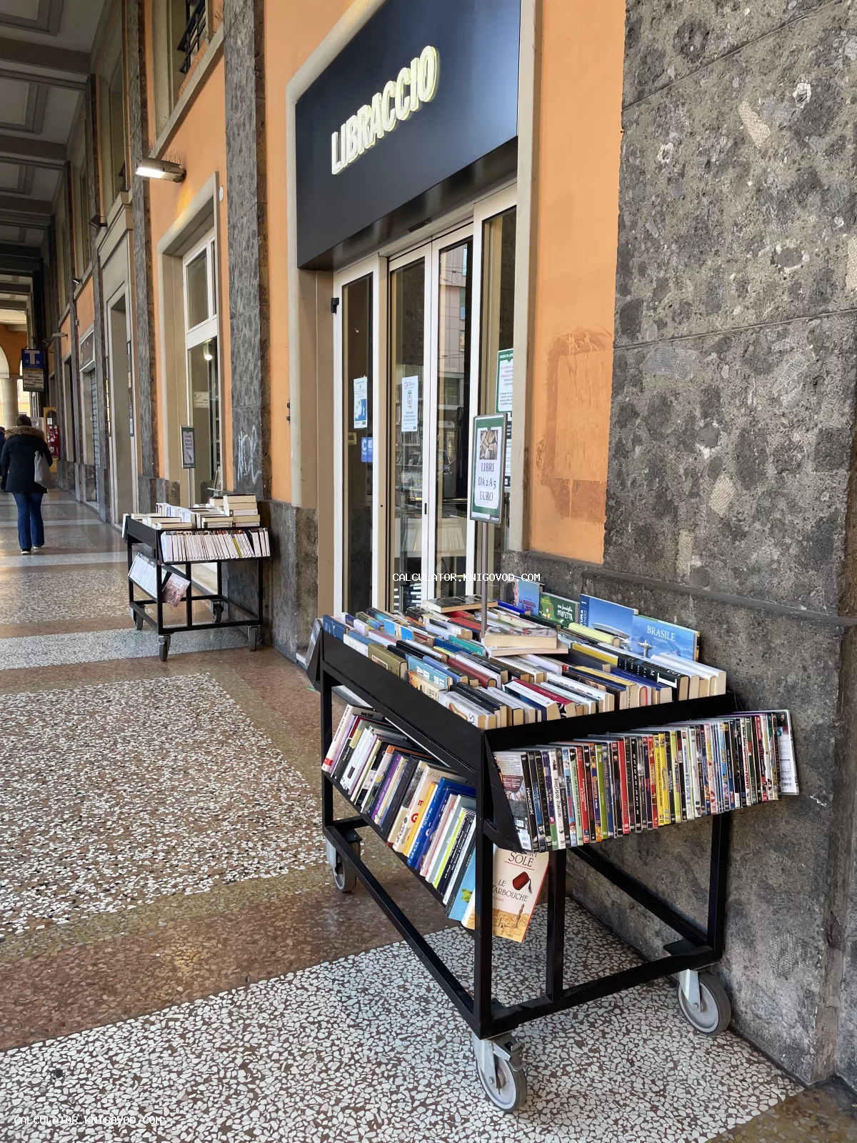 Мобильные стеллажи с книгами и медиа-изданиями перед входом в книжный магазин Libraccio под каменными сводами.