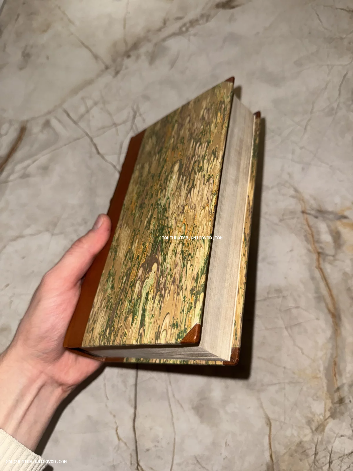 Антикварная книга с обложкой из мраморной бумаги и кожаными вставками в руке на светлом фоне.