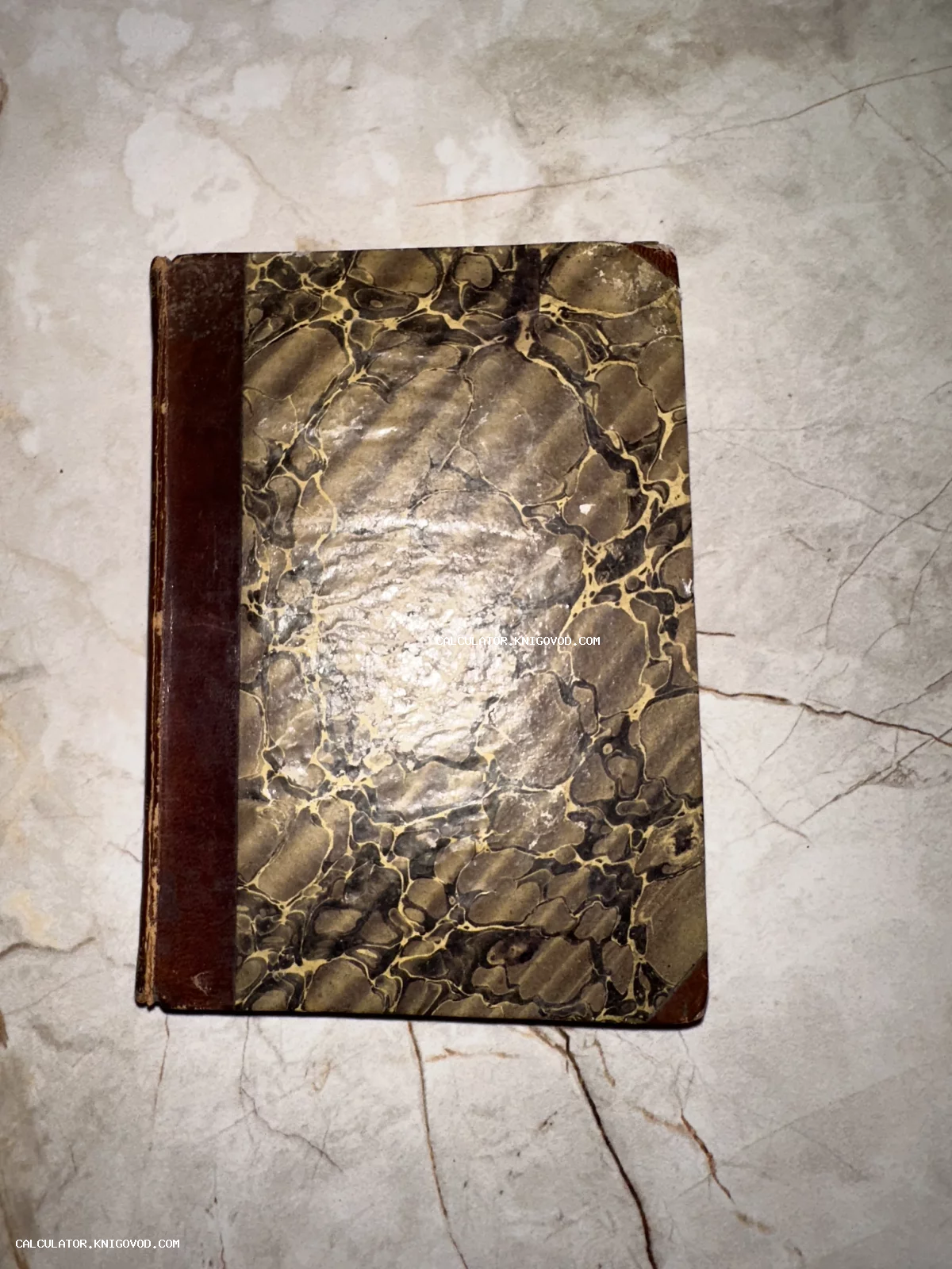 Старинная антикварная книга с темным корешком и обложкой, имитирующей рисунок мрамора, на светлой поверхности.