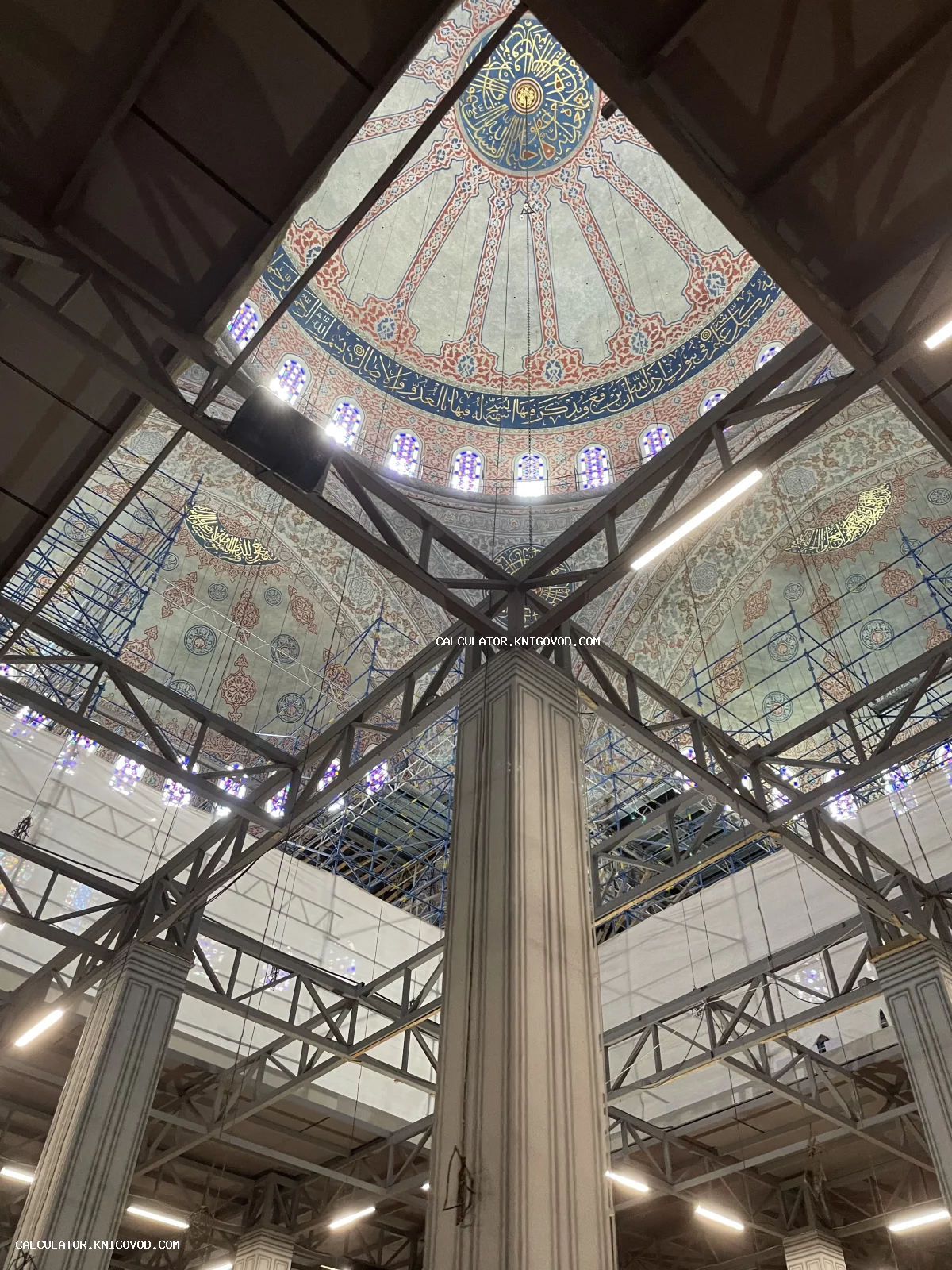 Вид снизу вверх на расписной купол мечети с каллиграфией, окруженный металлическими строительными лесами.