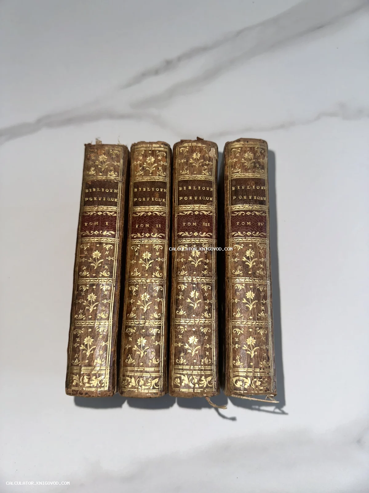 Четыре старинных тома в кожаных переплетах с богатым золотым тиснением на корешках, расположенные на светлой поверхности.