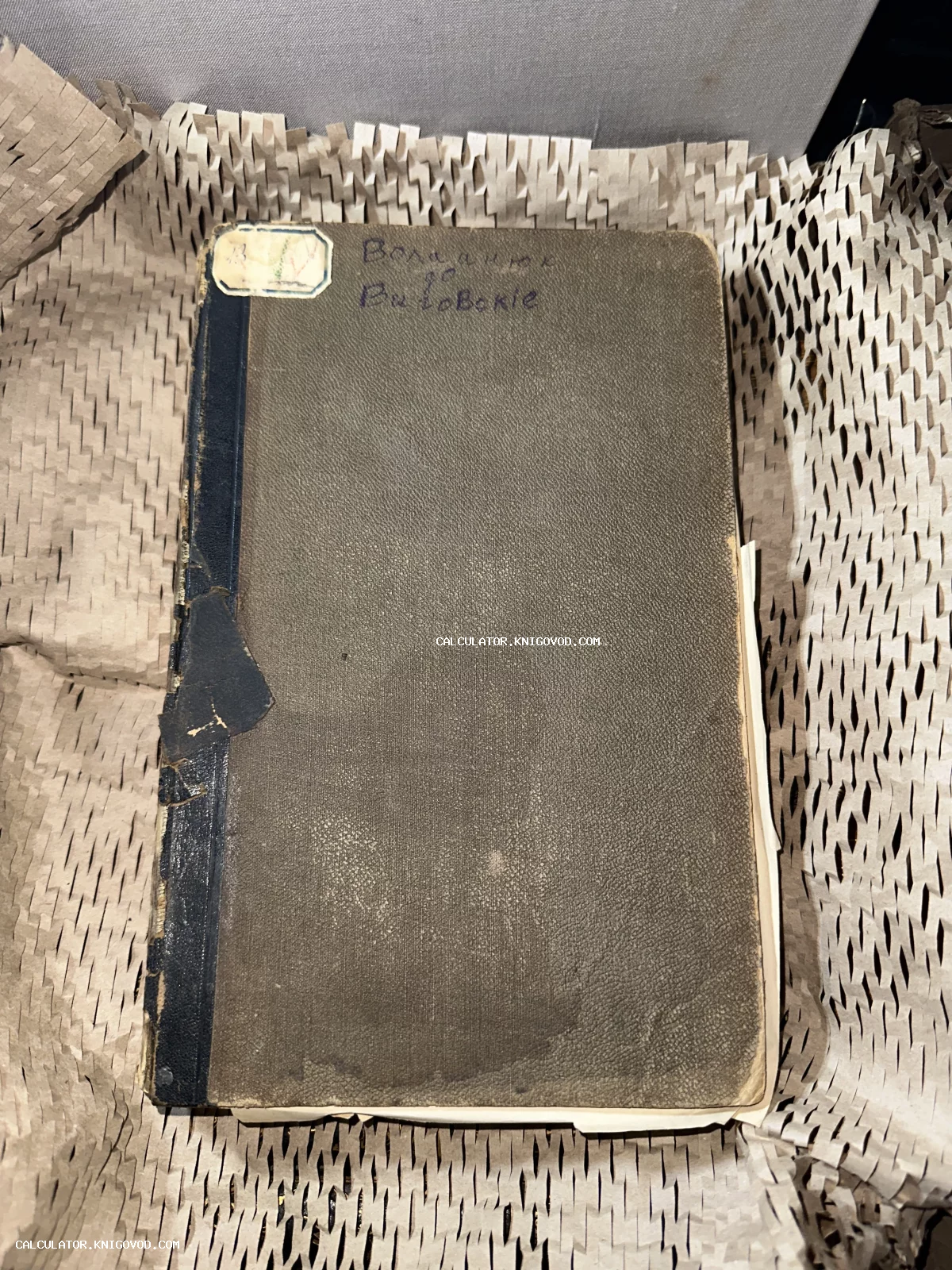 Вид сверху на старинную книгу в сером твердом переплете с темным корешком, рукописной надписью и архивным ярлыком на обложке.