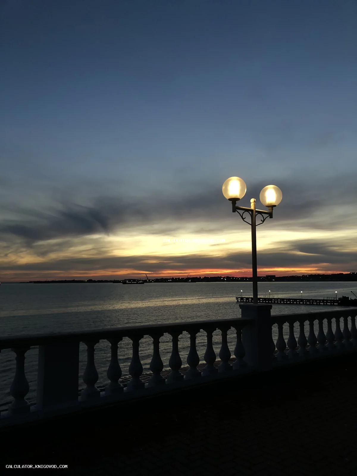 Уличный фонарь с двумя светящимися плафонами на фоне морского заката и каменной балюстрады.