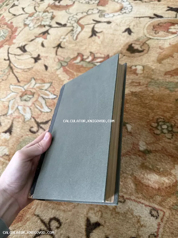 Рука держит закрытую антикварную книгу в серо-зеленом тканевом переплете на фоне ковра с цветочным узором.