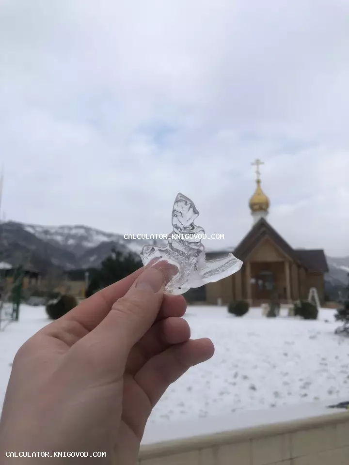 Рука держит прозрачный осколок льда в форме ангела на фоне деревянной церкви и заснеженных гор.