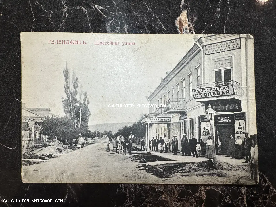 Черно-белая дореволюционная открытка с видом на Шоссейную улицу в Геленджике, изображающая здание Центральной столовой, вывески магазинов и людей.
